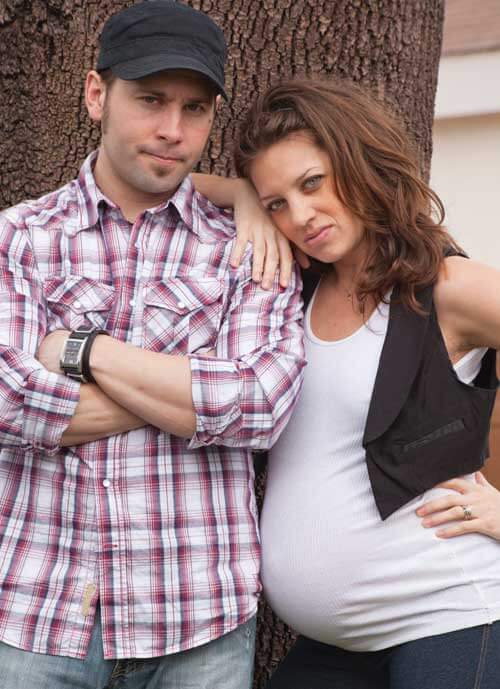 A pregnant women leaning on her partner. His arms are crossed. They are in front of a tree.