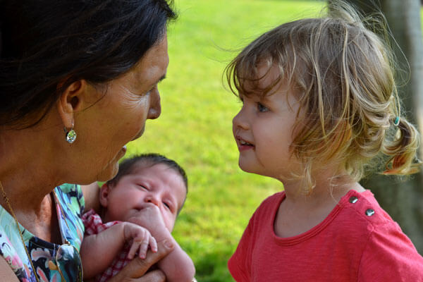 A toddler looking closely at an elderly women who is holding a baby